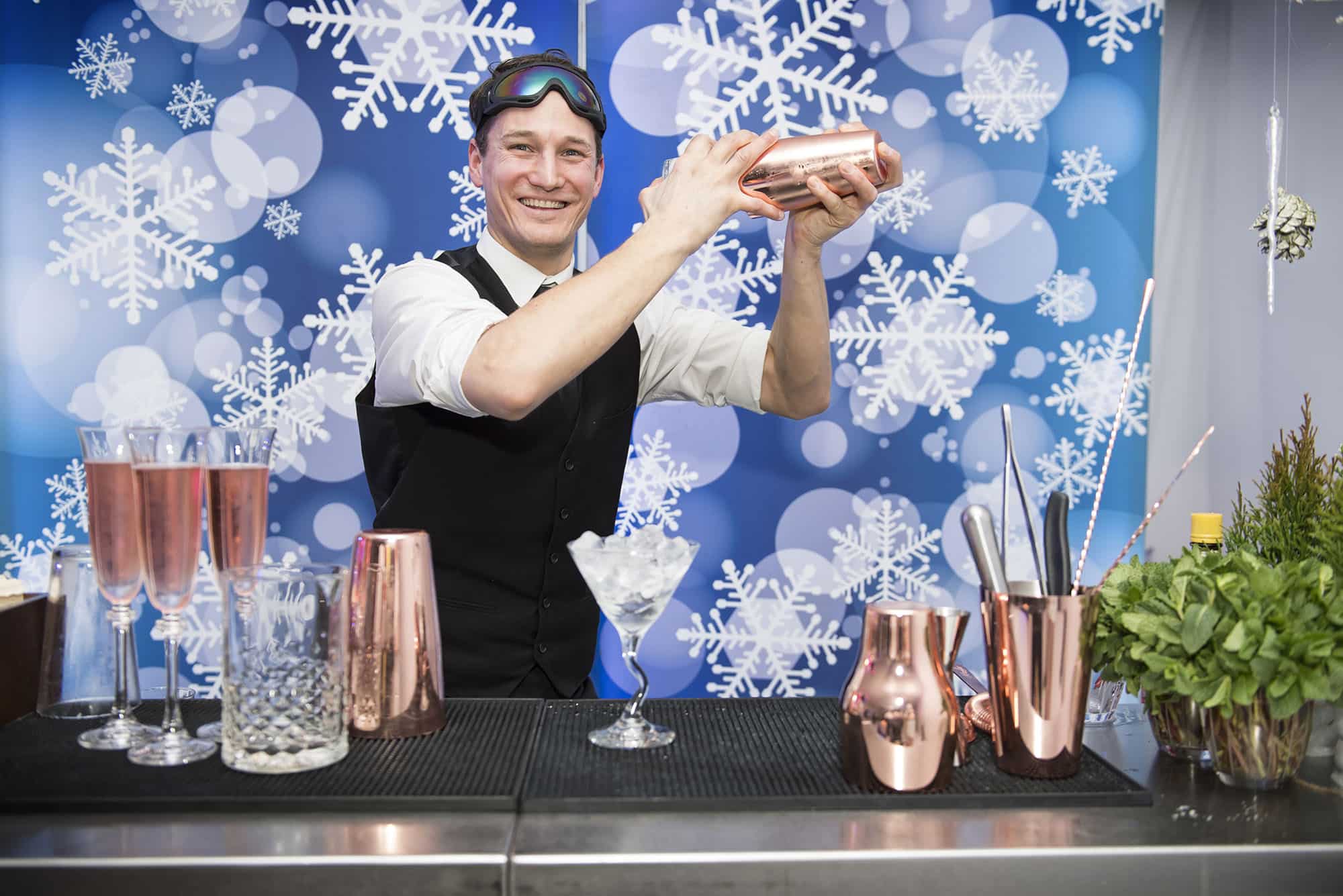 A barman shaking a cocktail behind a bar with a snowflake backdrop catering for a winter party