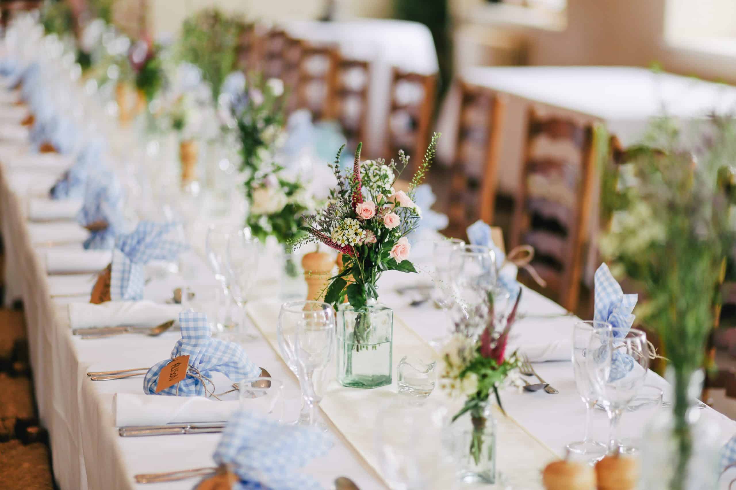 A row of long tables with chairs set out for a wedding breakfast. The table is decorated with napkins, plates and floral centres at every place setting