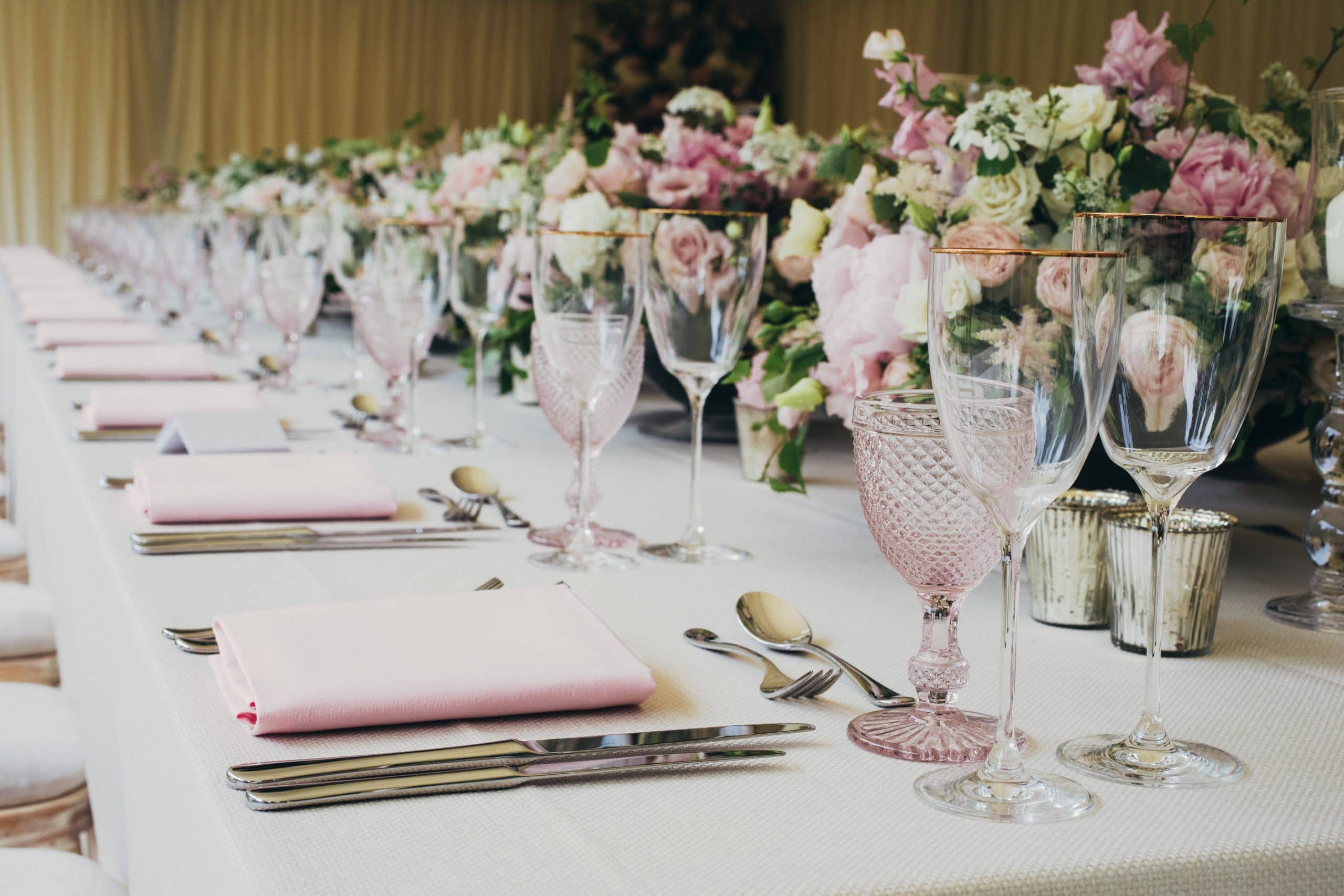 A table set for a wedding breakfast with white tablecloth, cutlery, napkins, glassware and colourful floral decorations along the length of the table. Wedding catering budget